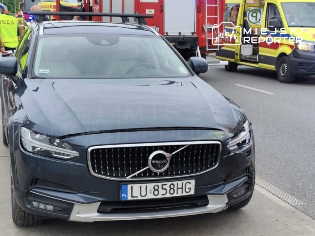 Czarny samochód marki Volvo zaparkowany obok wozu strażackiego i ambulansu, w tle pracujący funkcjonariusze.