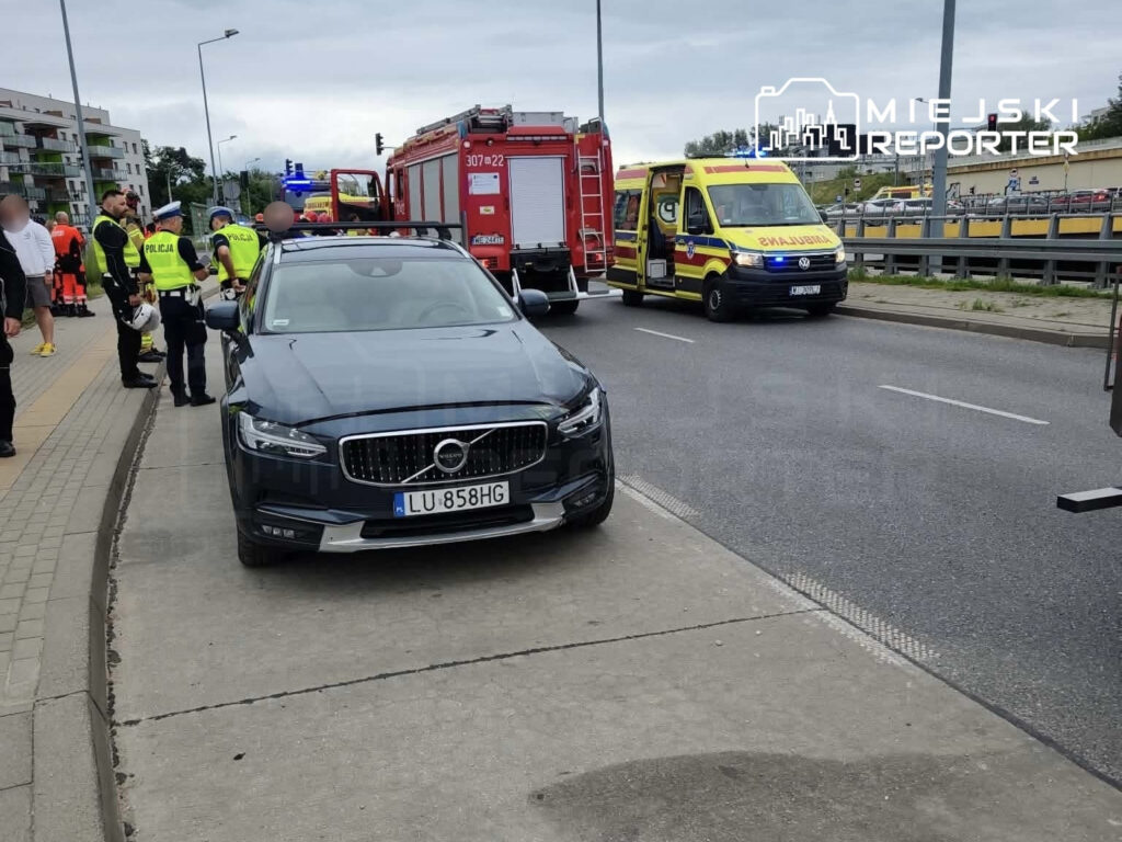 Na poboczu drogi stoją funkcjonariusze policji oraz strażacy, obok zaparkowany samochód marki Volvo. W tle widoczna karetka pogotowia.