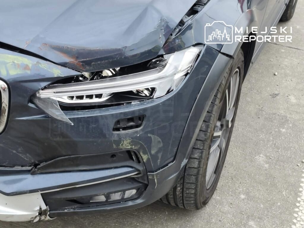 Uszkodzony samochód osobowy z wgniecioną maską i uszkodzoną lampą przednią na parkingu.