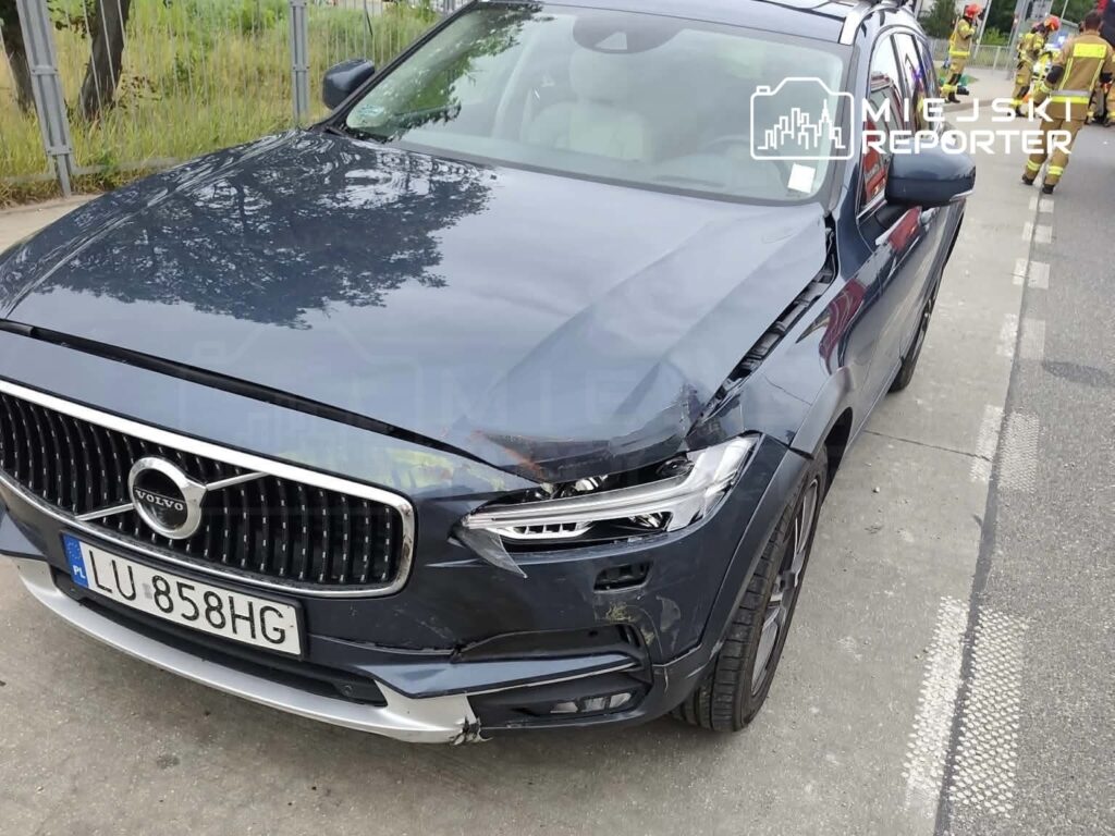 Uszkodzony samochód osobowy marki Volvo zaparkowany na poboczu drogi, obok widoczni strażacy.