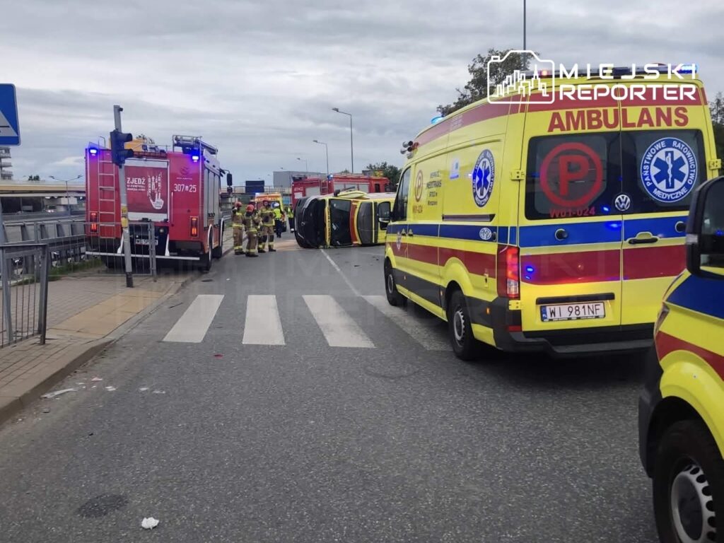 Na drodze leży przewrócone auto, a obok działają strażacy i karetki pogotowia w akcji ratunkowej.