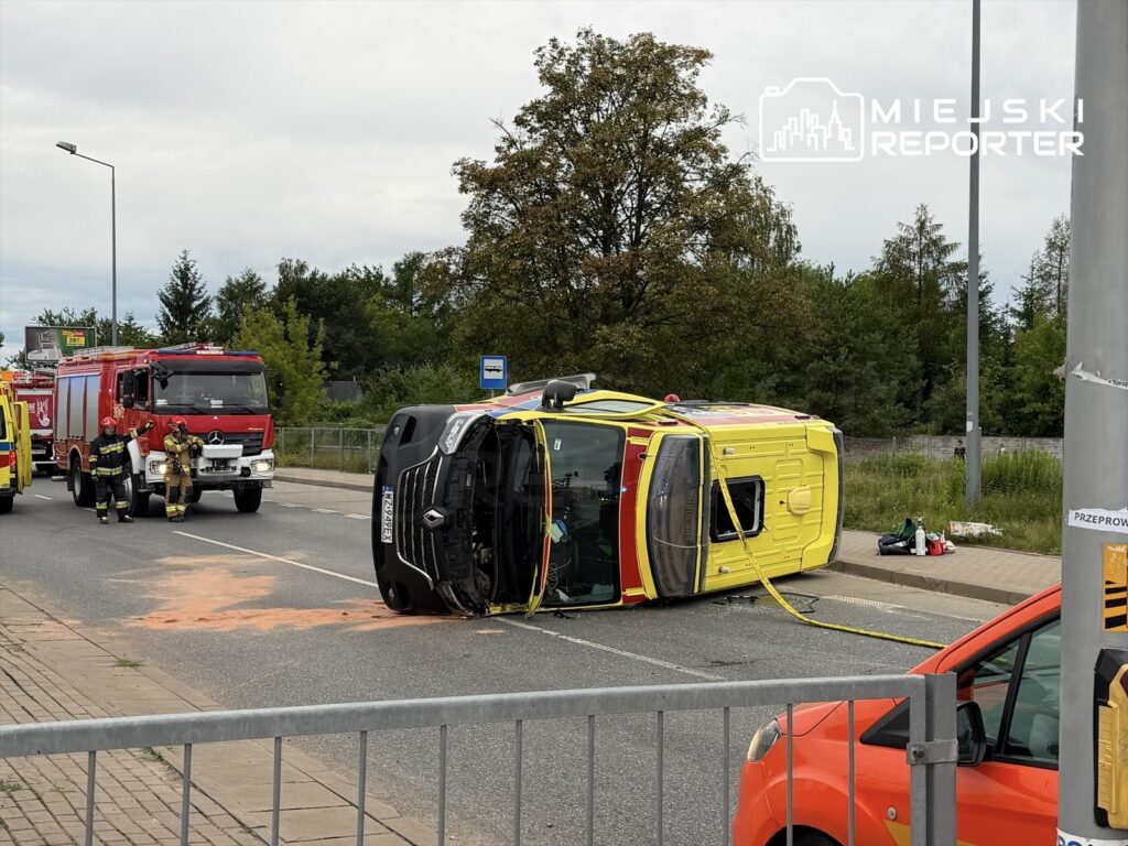 Na jezdni leży przewrócona karetka, a obok niej pracują strażacy w mundurach. W tle widoczne są dwa wozy strażackie.