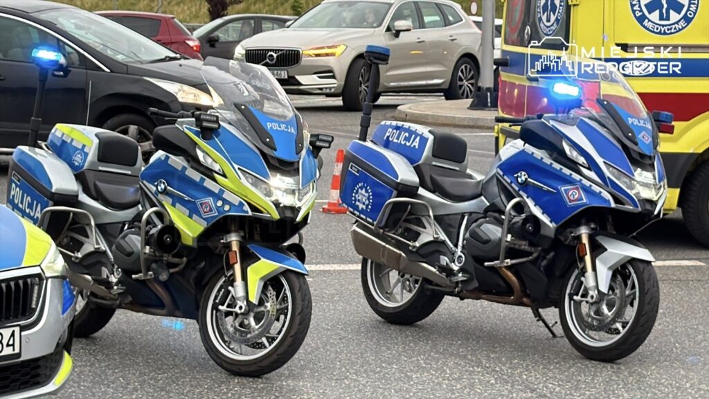 Dwa motocykle policyjne zaparkowane na jezdni w pobliżu karetki pogotowia, z włączonymi sygnałami świetlnymi.