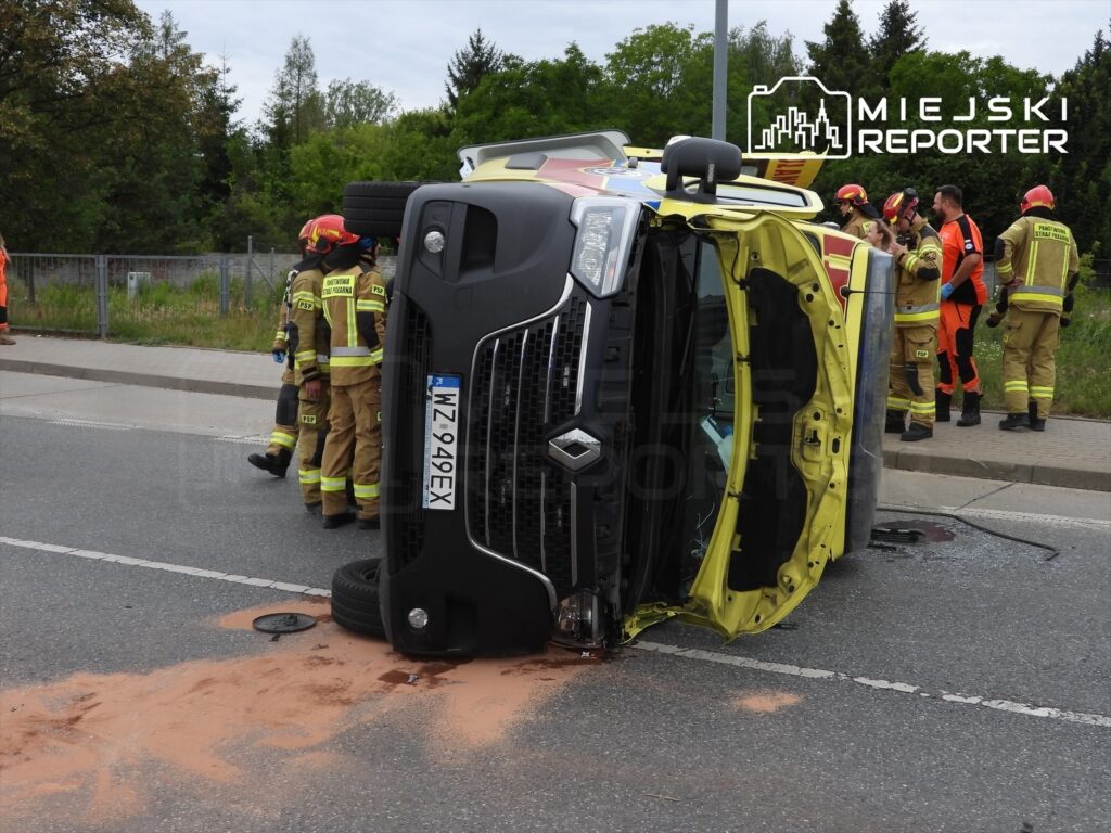 W przewróconej karetkę na drodze, obok grupy strażaków i ratowników medycznych, widoczne plamy płynów.
