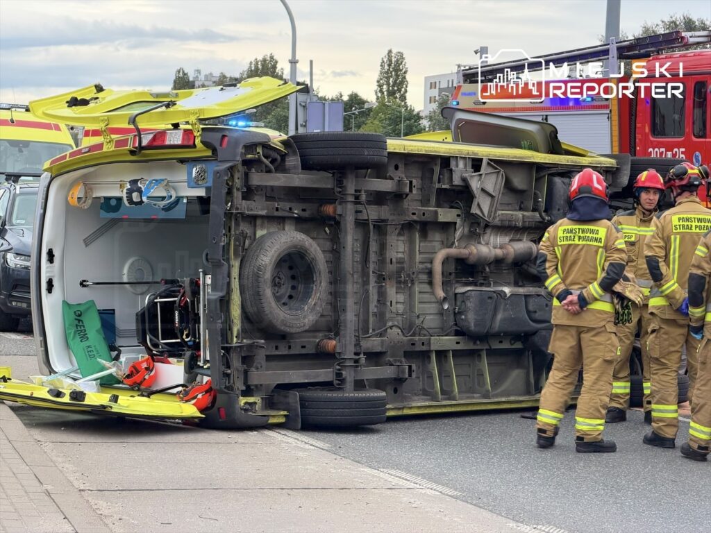 Ambulans przewrócony na bok po zderzeniu z innym pojazdem, otoczony przez strażaków w mundurach, na ruchliwej ulicy.