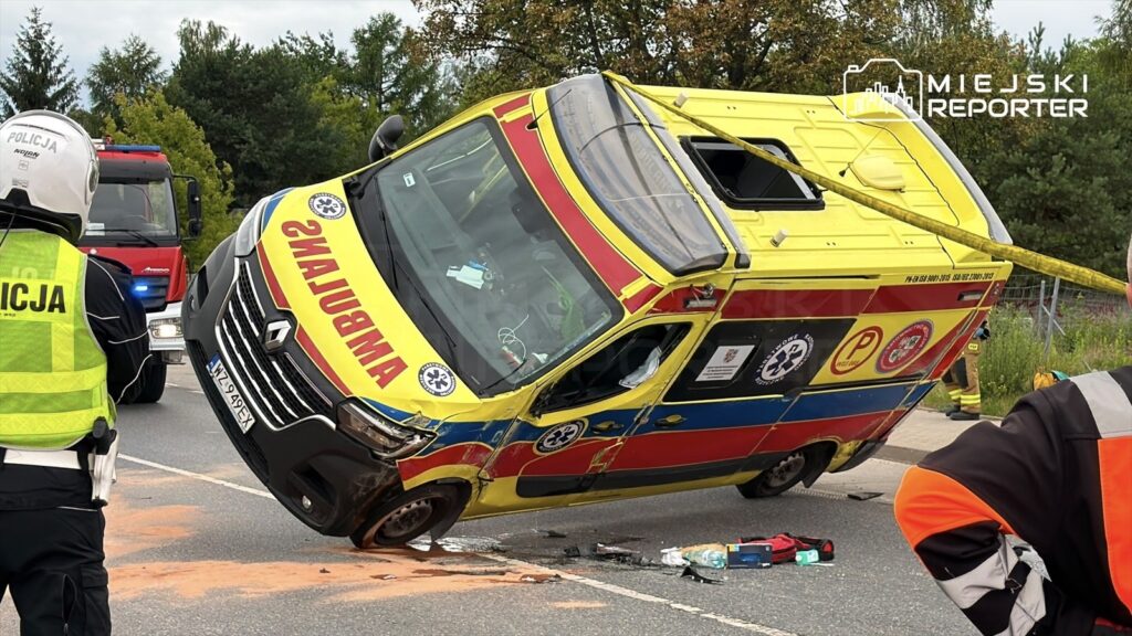 Ambulans przewrócony na bok po zderzeniu, obok stojący policjant w kamizelce odblaskowej, w tle woz strażacki.
