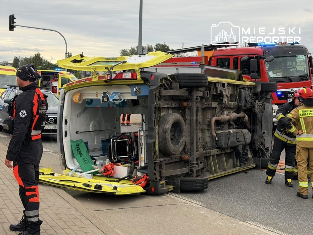 Na drodze leży przewrócona karetka, obok grupka ratowników i strażaków w akcji. W tle widoczne inne pojazdy ratunkowe.