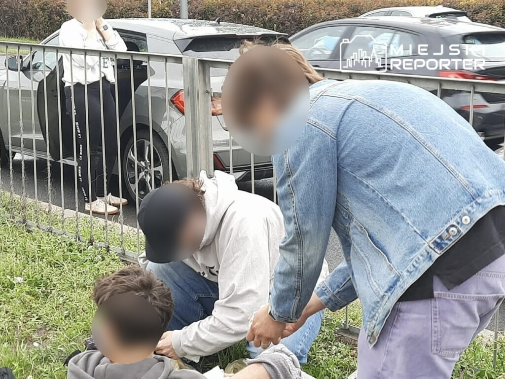 Młody mężczyzna w bluzie pomaga poszkodowanemu siedzącemu na trawie obok samochodu, podczas gdy kolejna osoba obserwuje z tyłu.