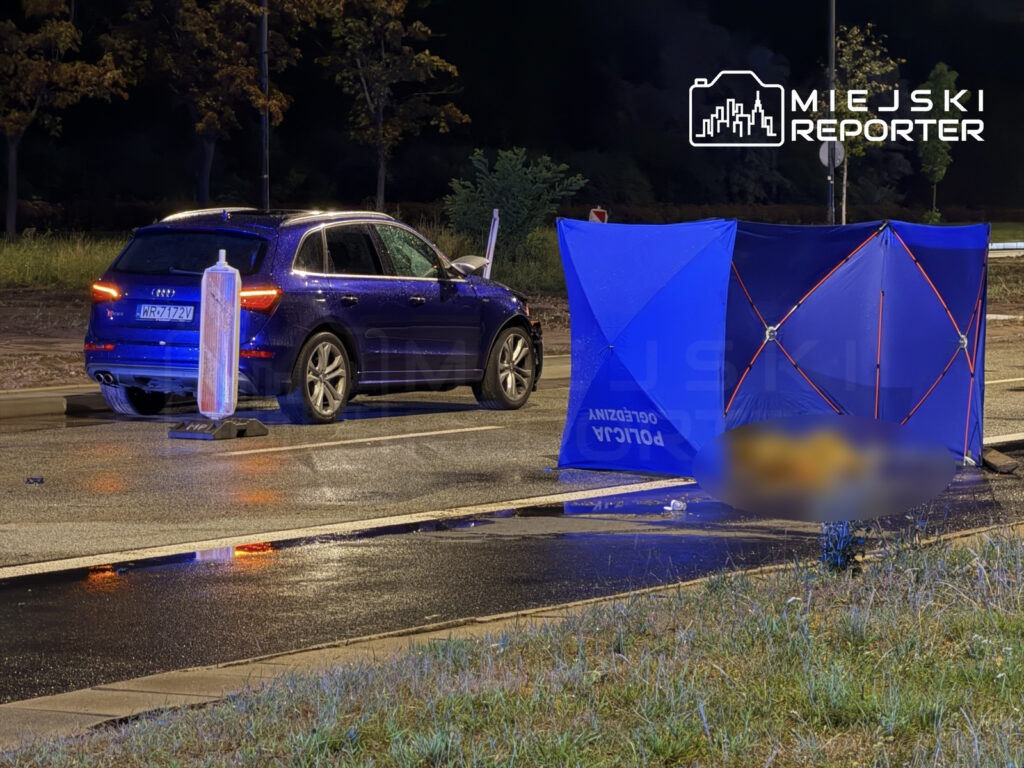 Audi Q5 zatrzymane na drodze, obok niebieski namiot policyjny oraz znak ostrzegawczy, mokra nawierzchnia po deszczu.