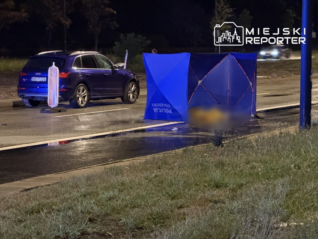 Na jezdni stoi niebieski namiot policyjny, obok zaparkowany ciemny SUV. W tle widoczny jest mokry asfalt po deszczu.