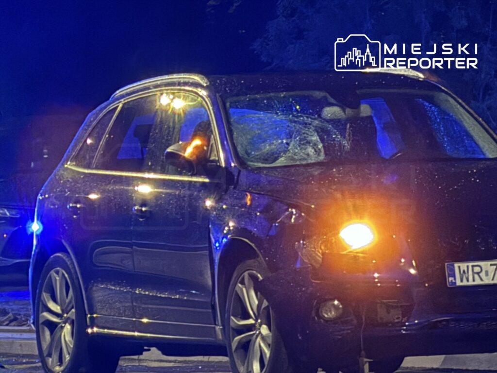 Uszkodzony czarny SUV z rozbitą szybą, zaparkowany w oświetlonym niebieskim świetle policyjnym.
