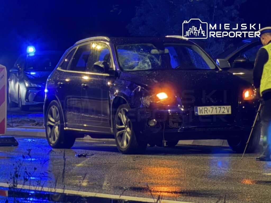 Czarny SUV z uszkodzoną przednią częścią stoi na mokrej nawierzchni, otoczony przez policyjne pojazdy z niebieskimi światłami.