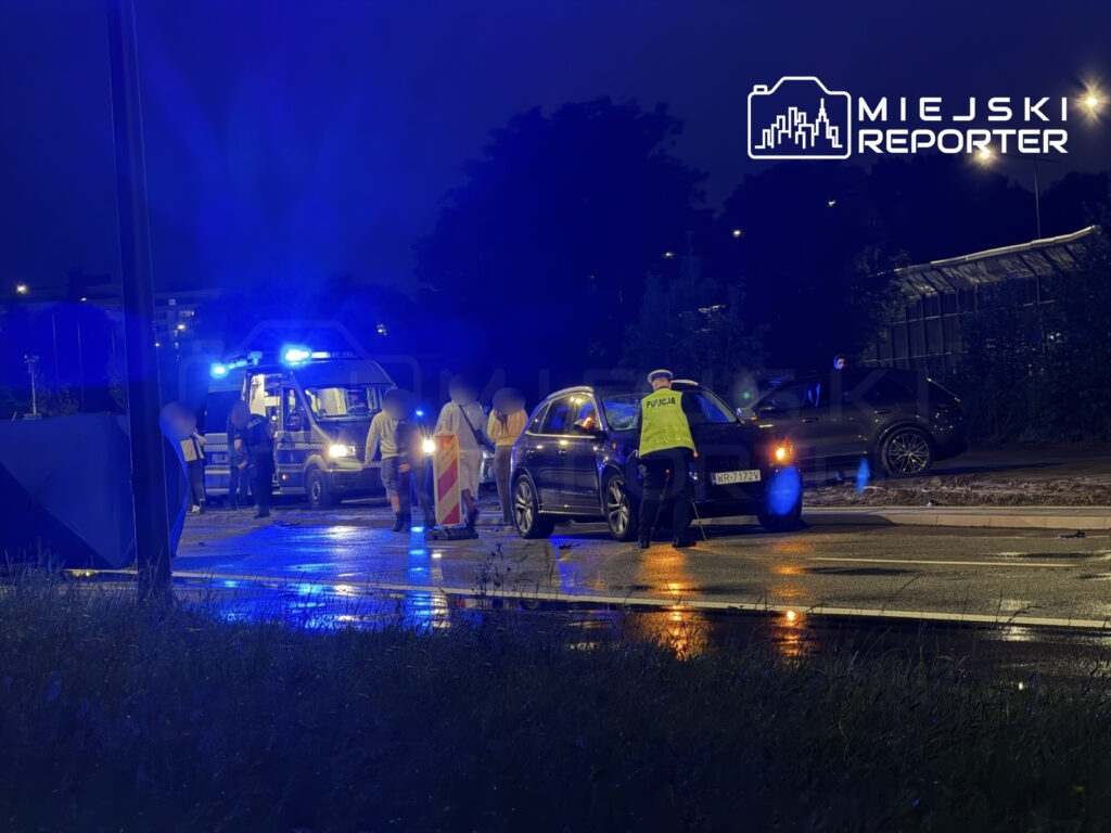 Grupa policjantów przeprowadza kontrolę drogową w nocy, obok radiowozu z włączonymi sygnalizatorami świetlnymi.