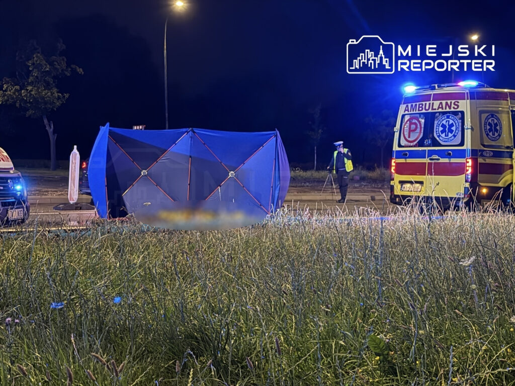 Na drodze leży przewrócony pojazd, a obok pracuje policjant oraz karetka pogotowia z włączonymi sygnałami świetlnymi.