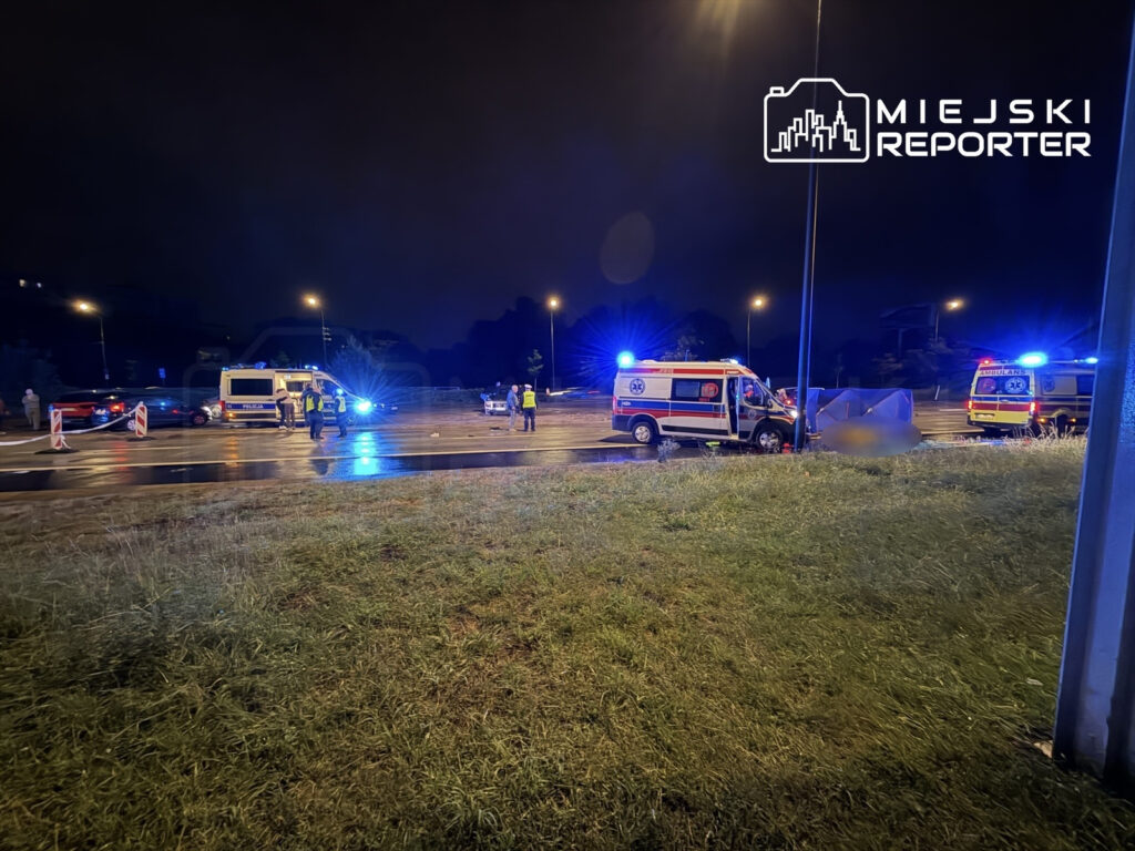 Na drodze w nocy zderzyły się dwa samochody, w tle widoczne karetki i policja zabezpieczająca miejsce zdarzenia.