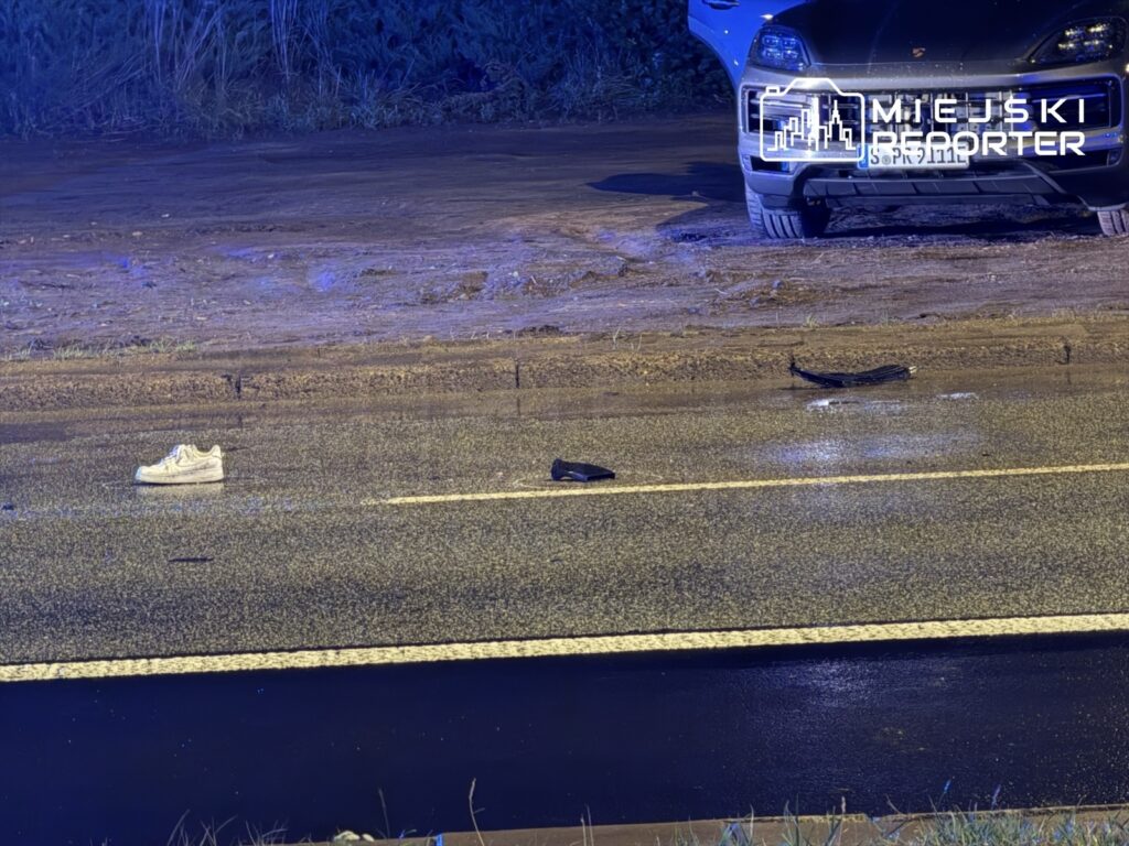 Na mokrej nawierzchni drogi leżą porzucone buty – biały sneaker i czarny sandał, obok stoi czarny samochód.