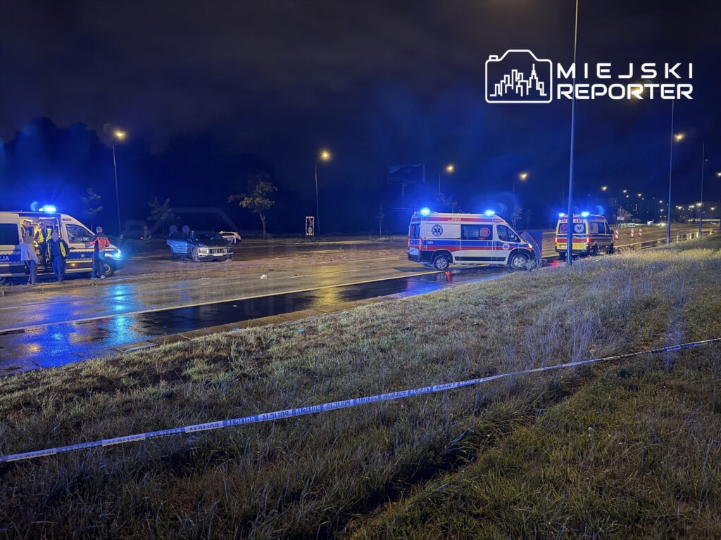 Służby ratunkowe obsługujące wypadek na mokrej drodze, z niebieskimi światłami na ambulanse i policyjny radiowóz.