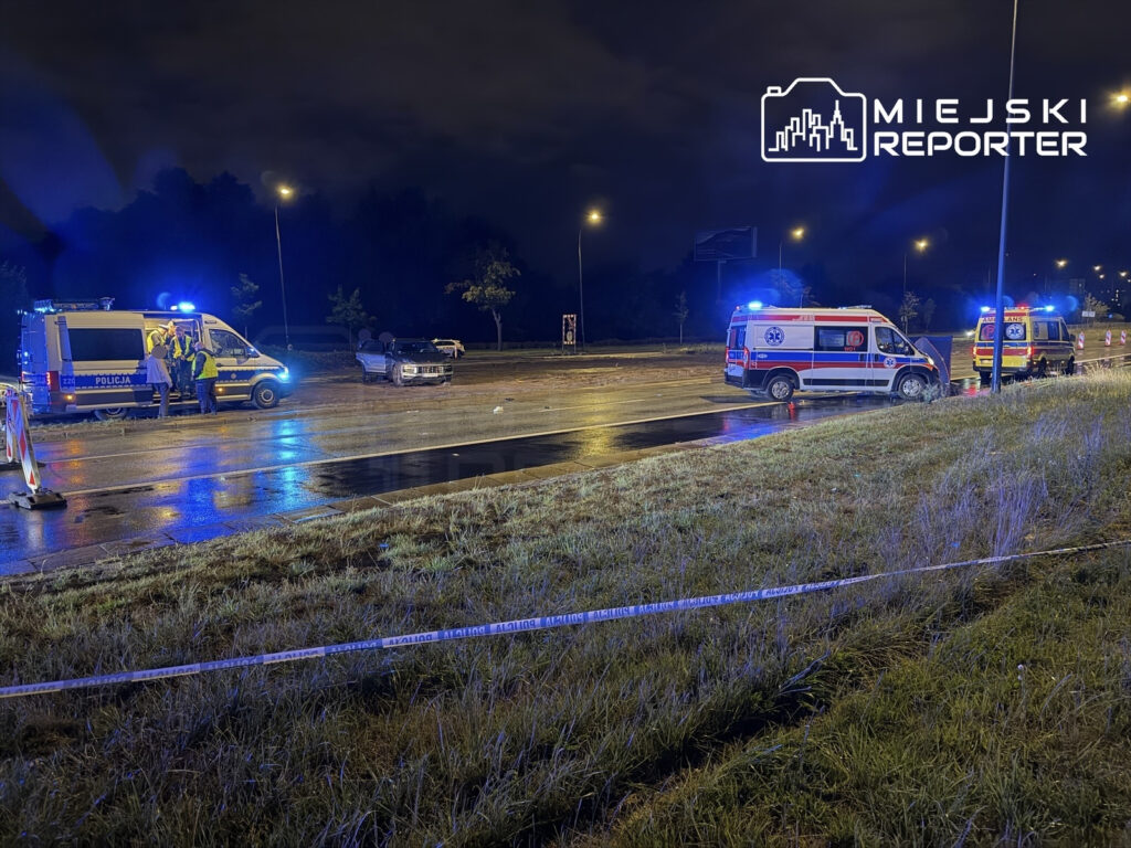 Na drodze, w nocy, kilka pojazdów ratunkowych z włączonymi sygnałami świetlnymi oraz policjanci prowadzący działania na miejscu zdarzenia.