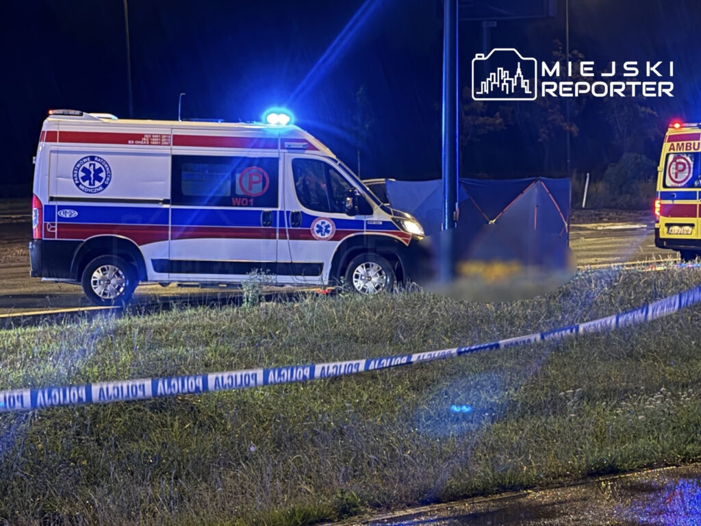 Ambulans z włączonymi sygnałami świetlnymi stoi na poboczu drogi, wokół widoczne taśmy policyjne i namiot osłonowy.
