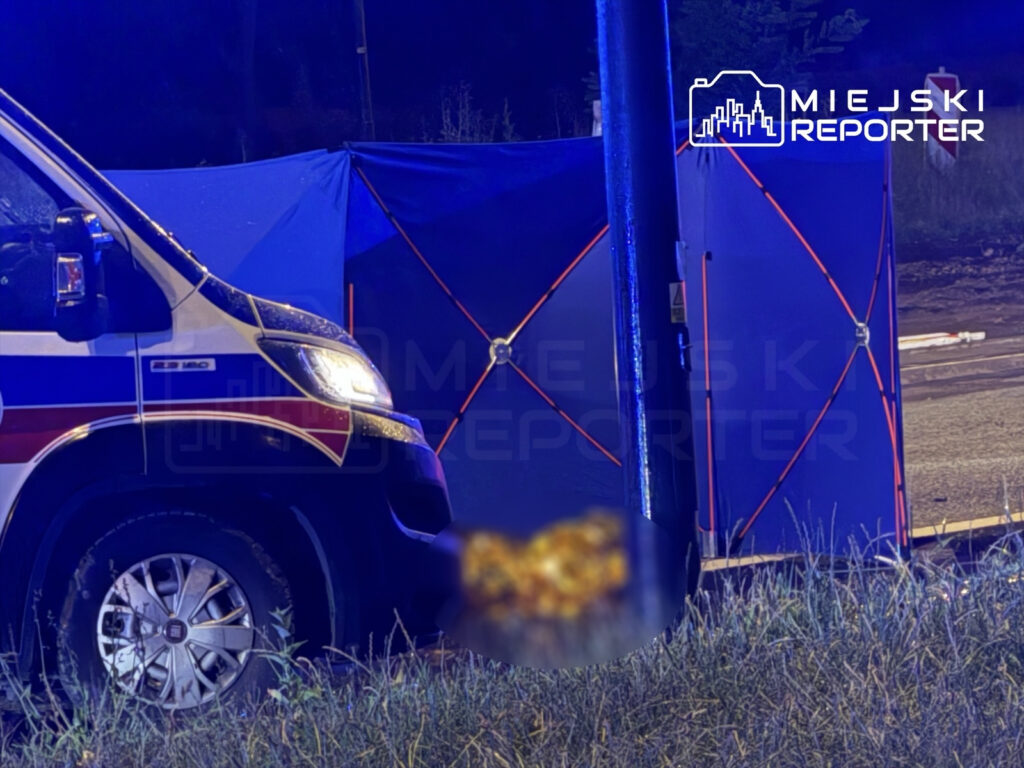 Ambulans z włączonymi światłami stoi obok niebieskiego namiotu, w otoczeniu trawy i drogi.