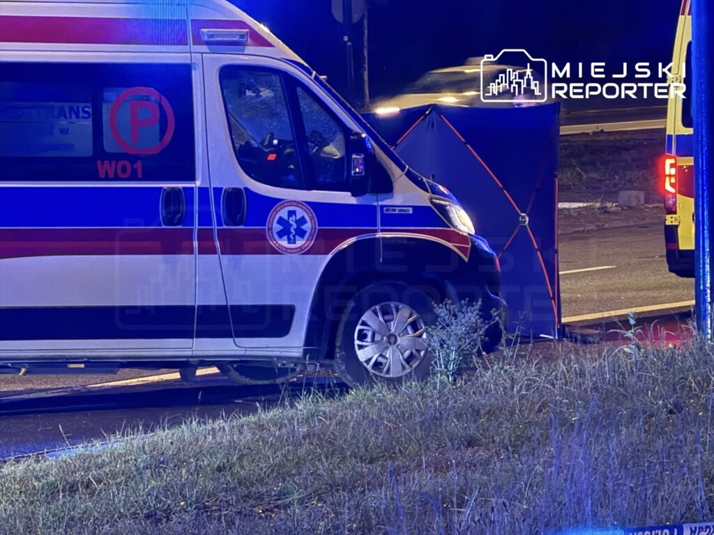 Karetka pogotowia zatrzymana na drodze, z niebieskimi światłami w nocy, obok osłony zabezpieczającej miejsce zdarzenia.