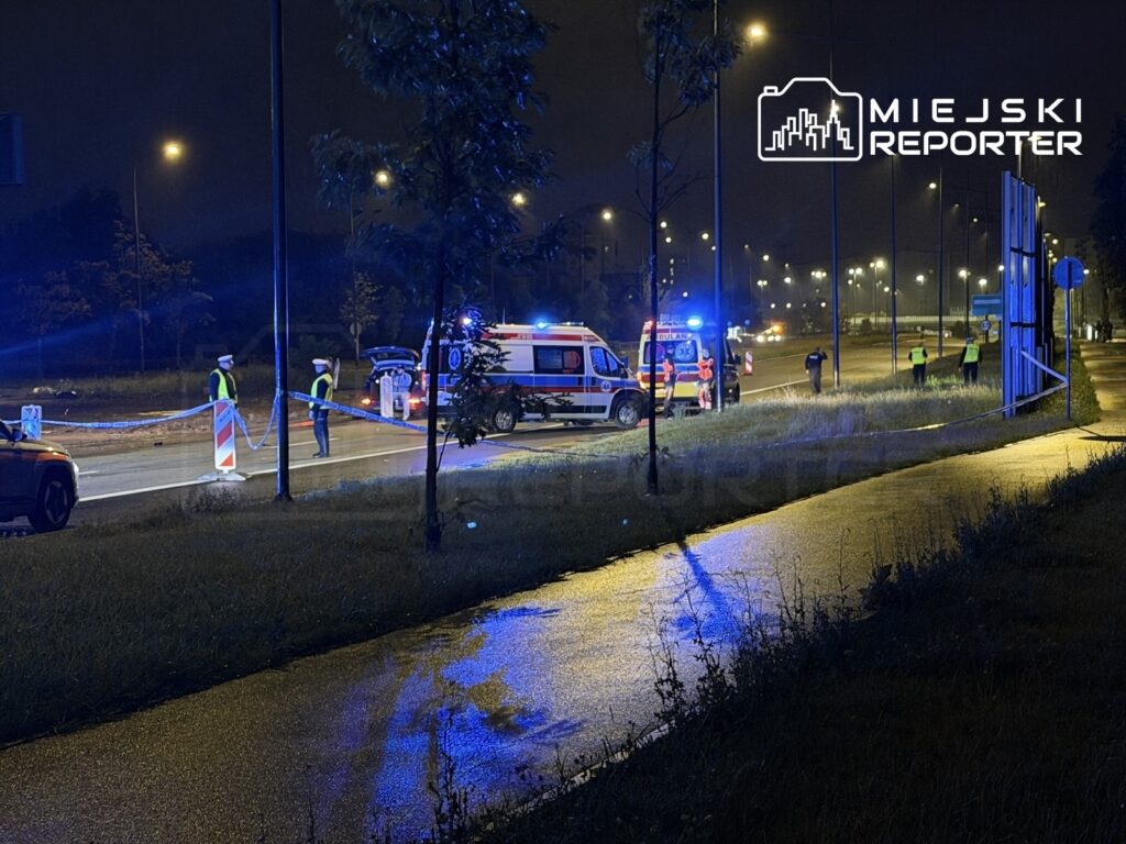 Na drodze nocą, policjanci zabezpieczają miejsce zdarzenia, a w tle widać ambulans i światła policyjne.