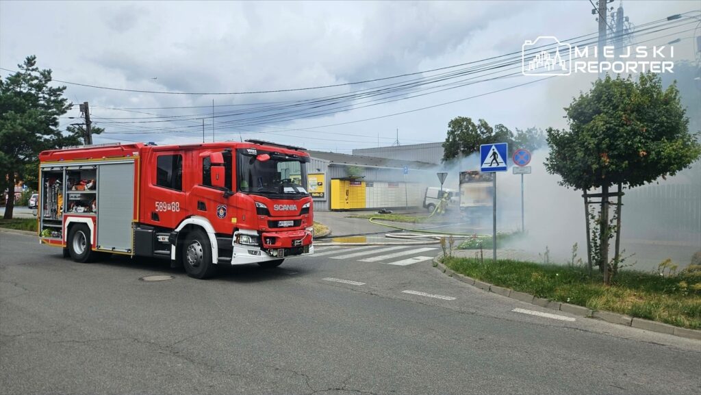 Czerwony wóz strażacki zatrzymany przy skrzyżowaniu, w tle widoczny dym wydobywający się z parkingu.