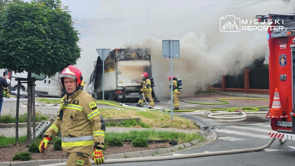 Strażak w hełmie i mundurze gaśniczym przemieszcza się obok palącego się busa, z którego wydobywa się dym.