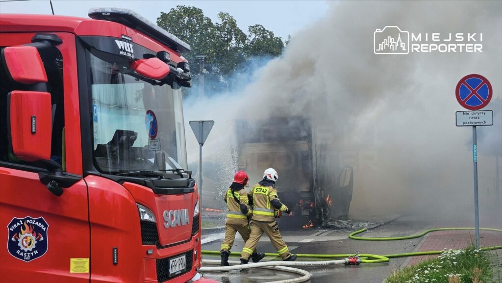 Strażacy w mundurach gaśniczych podchodzą do płonącego busa na parkingu, obok widać wóz strażacki.