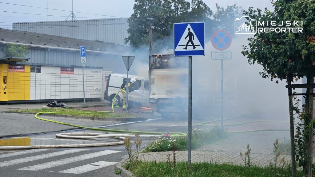 Strażak w mundurze gaśniczym polewa wodą palącego się busa na parkingu, w tle widoczne znaki drogowe i dym.