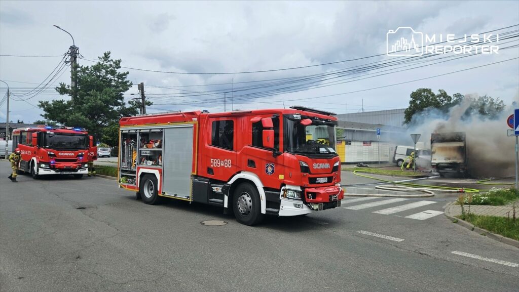 Czerwony wóz strażacki z włączonymi sygnałami świetlnymi podjeżdża do miejsca pożaru, gdzie dymi uszkodzony bus.