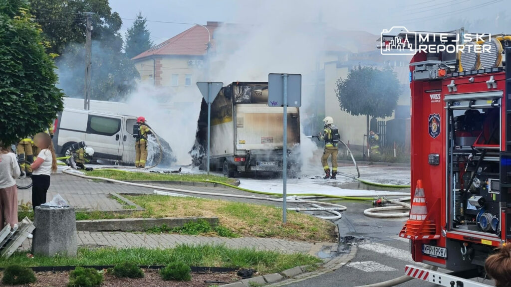 Strażacy gaszą pożar busów na parkingu, obok widać węże strażackie oraz dym unoszący się w powietrzu.