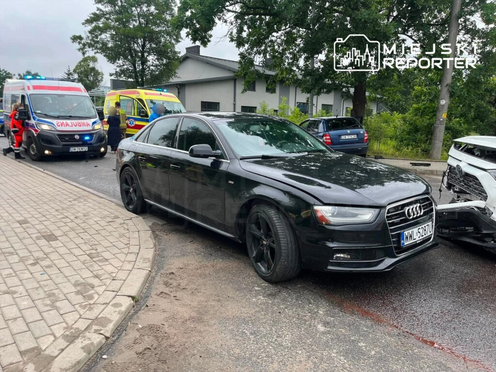 Czarny samochód marki Audi stoi na skrzyżowaniu, obok dwóch ambulansów z włączonymi sygnałami świetlnymi.