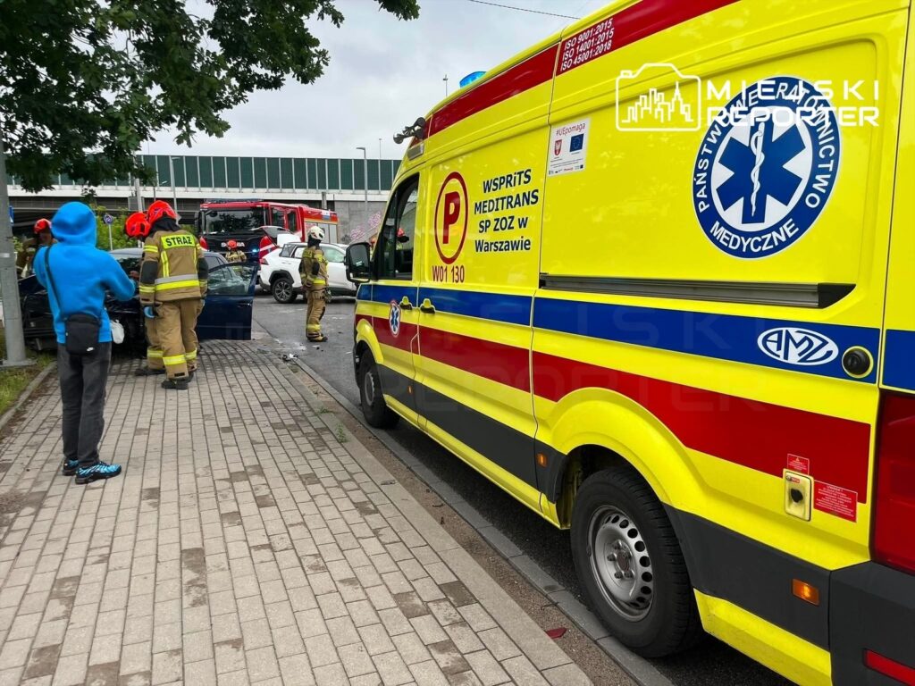 Ratownicy medyczni i strażacy udzielają pomocy poszkodowanym w wypadku drogowym, obok uszkodzonych pojazdów.