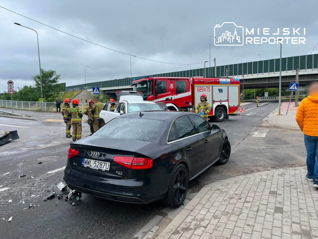 Czterech strażaków w mundurach i kaskach bada miejsce wypadku na skrzyżowaniu, obok uszkodzonego Audi.