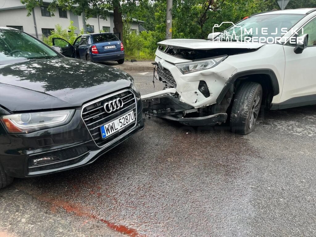 Czarny samochód marki Audi z uszkodzoną maską zderza się z białym SUV-em, który ma zniszczony przód, na ulicy pokrytej wodą.