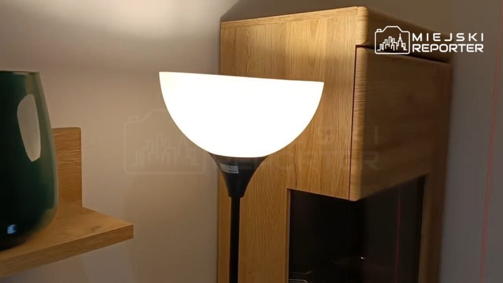 Lampa podłogowa z okrągłym kloszem o białym świetle stoi obok drewnianej komody w nowoczesnym wnętrzu.