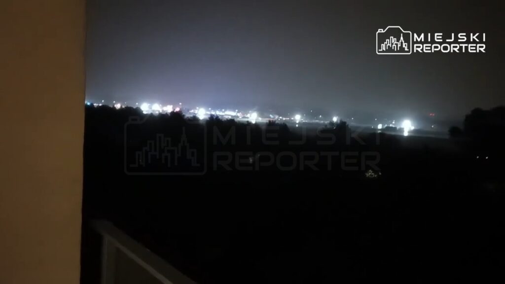 Widok na oświetloną okolicę nocą, z migoczącymi światłami w tle, w atmosferze zamglenia.