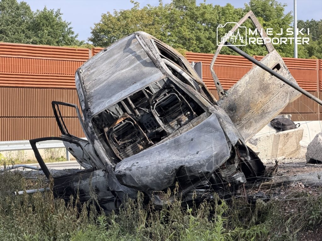 Spalony pojazd leży na poboczu drogi, otoczony wysoką trawą, z widocznymi zniszczeniami wnętrza.