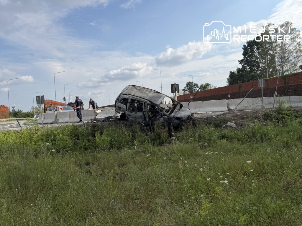 W pobliżu drogi leży uszkodzony samochód, obok którego policjanci prowadzą czynności dochodzeniowe.