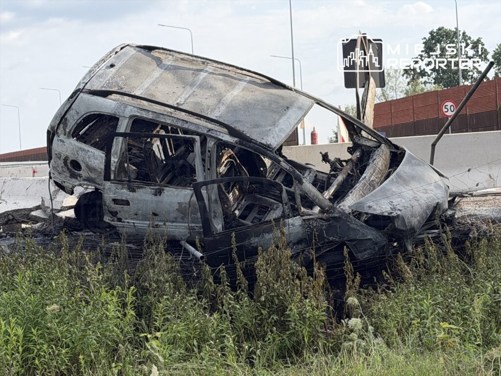 Zniszczony samochód leży na boku przy drodze, otoczony wysoką trawą i zniszczonymi barierkami.