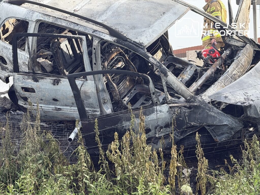 Zniszczony samochód osobowy leży na boku w zaroślach, a obok niego stoi strażak z narzędziem.