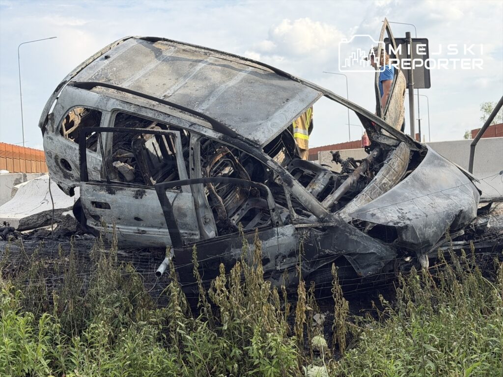 Zniszczony samochód leży na boku w zaroślach przy drodze, obok pracowników w kamizelkach odblaskowych.