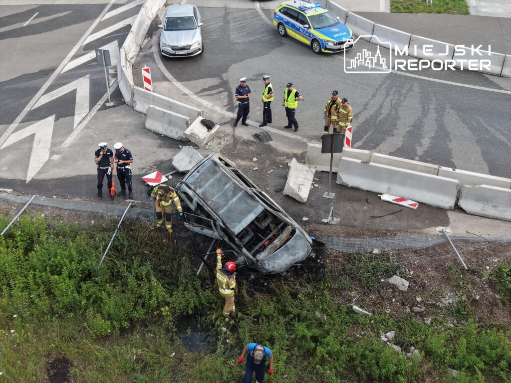 Grupa ratowników i policjantów pracuje przy wraku spalonego samochodu na poboczu drogi, obok ustawionych barier.
