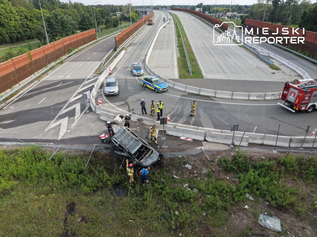 Na poboczu drogi leży spalone auto, wokół niego pracują strażacy i policjanci, a w tle widać ruch na jezdni.
