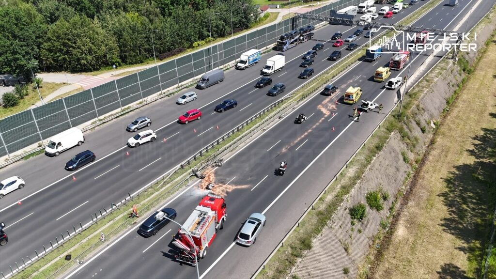 Wielu ratowników i pojazdów służb drogowych pracuje na pasie ruchu, gdzie leżą fragmenty silnika i kół.