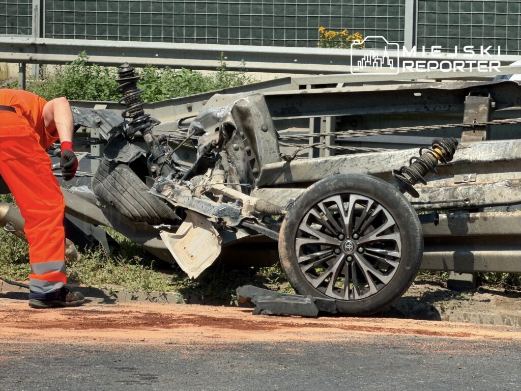 Mężczyzna w pomarańczowym uniformie naprawia uszkodzony pojazd na poboczu drogi, obok metalowej barierki.
