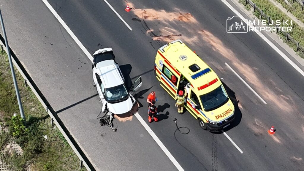 Na pasie ruchu stoi uszkodzona biała Toyota z otwartymi drzwiami, obok niej zespół ratunkowy rozmawia z kierowcą. W tle widać ambulans oraz znaki ostrzegawcze.