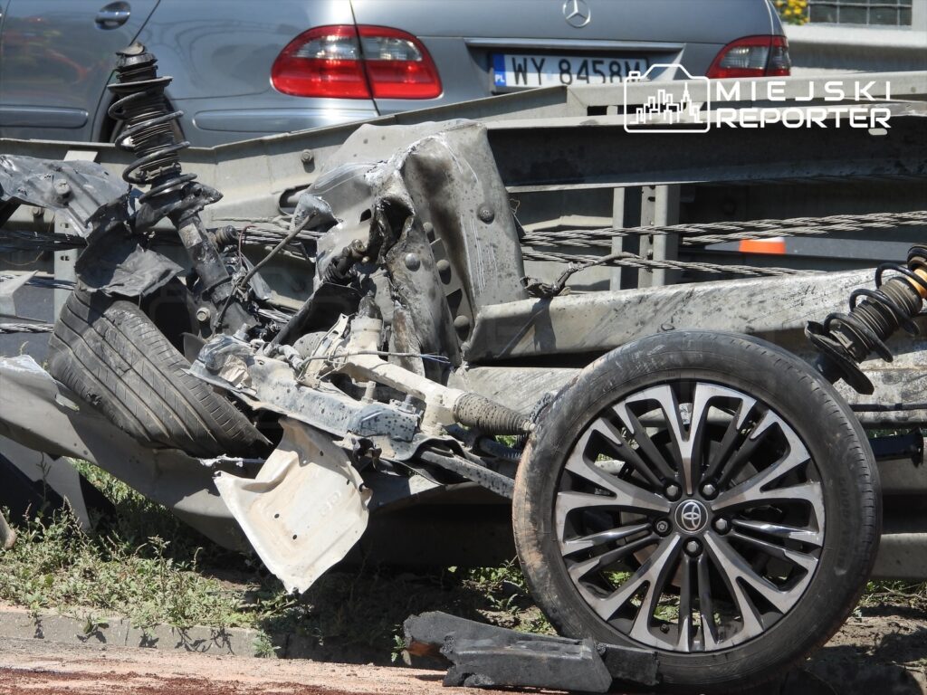 Zdemontowana część samochodu osobowego z widocznym uszkodzeniem zawieszenia i oponą leżącą na drodze.