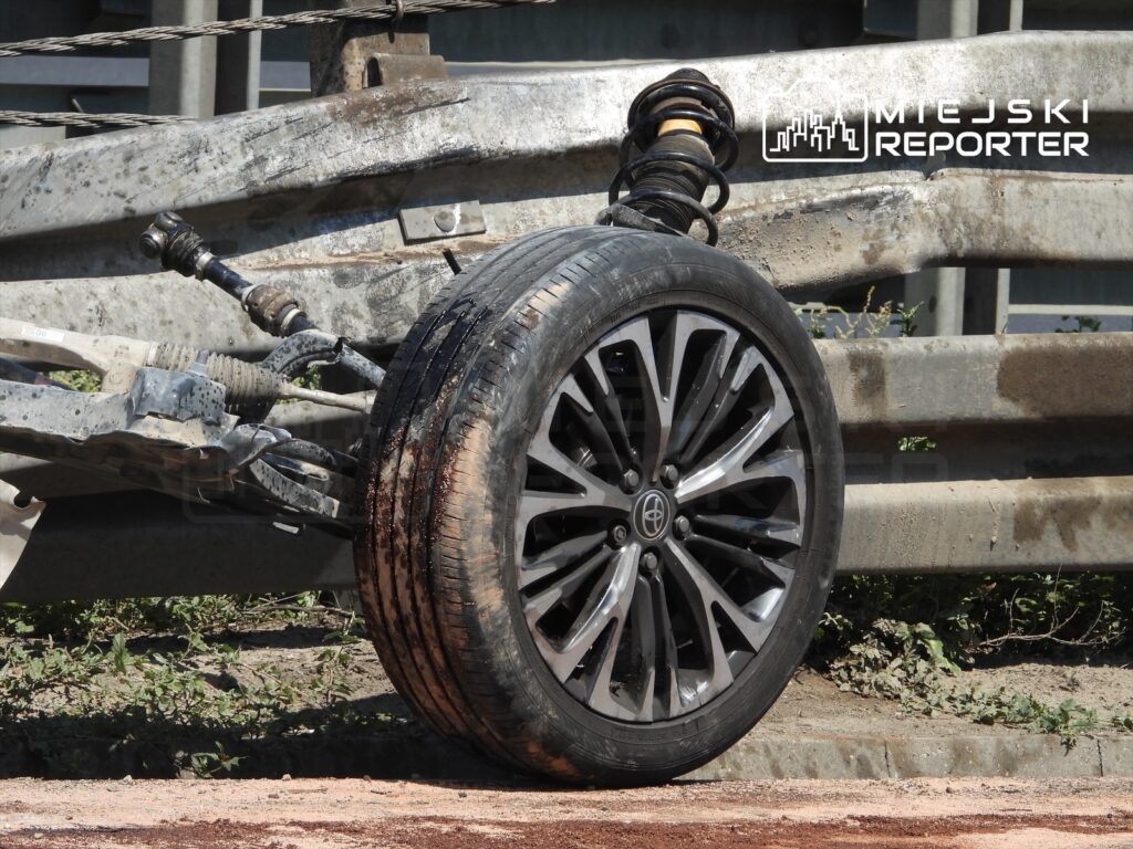 Zerwana opona leży obok uszkodzonego zawieszenia na poboczu drogi, w otoczeniu metalowych barier.